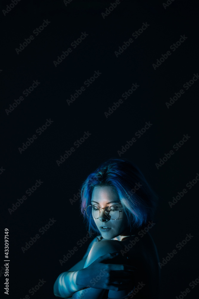 Female loneliness. Art portrait. Gender discrimination abuse. Tender insecure blue hair woman in eyeglasses embracing herself in neon light isolated on dark night copy space. Female loneliness. Art portrait. Gender discrimination abuse. Tender insecure blue hair woman in eyeglasses embracing herself in neon light isolated on dark night copy space.