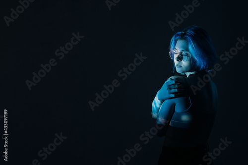 Body shaming. Art portrait. Self acceptance. Embarrassed shy imperfect woman in eyeglasses embracing herself in blue neon light isolated on dark night empty space background.