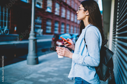 Side view of pensive hipster girl in casual wear thinking about touristic vacations during coffee break,thoughtful female blogger with smartphone device and caffeine beverage feeling pondering in city