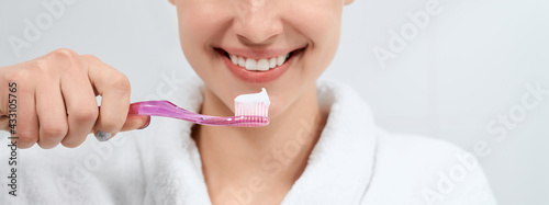 Close up of smiling young woman in white robe holding toothbrush with paste on light background. Concept of preparing for cleaning teeth at home with good mood.