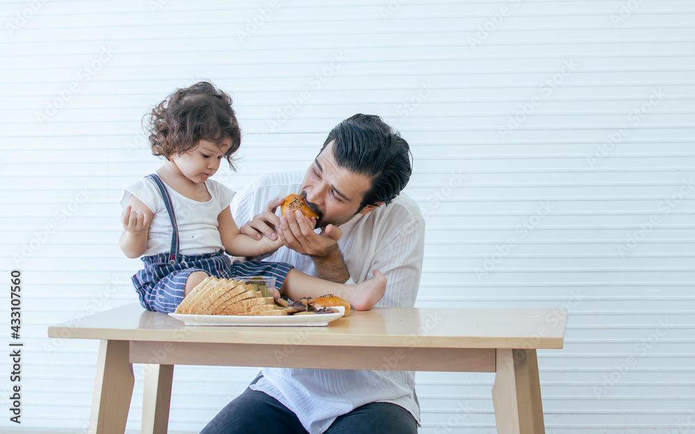 Caucasian handsome beard father or dad eating cookies and bread on the table at home together with his little adorable little girl. Copy space for advertisement. Education, Family and Kid Concept. Caucasian handsome beard father or dad eating cookies and bread on the table at home together with his little adorable little girl. Copy space for advertisement. Education, Family and Kid Concept.
