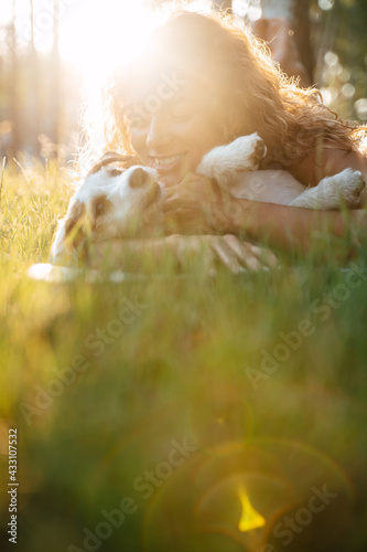 A woman with curly hair lies on the green grass with the dog jack russell in the meadow. The setting sun on the background.