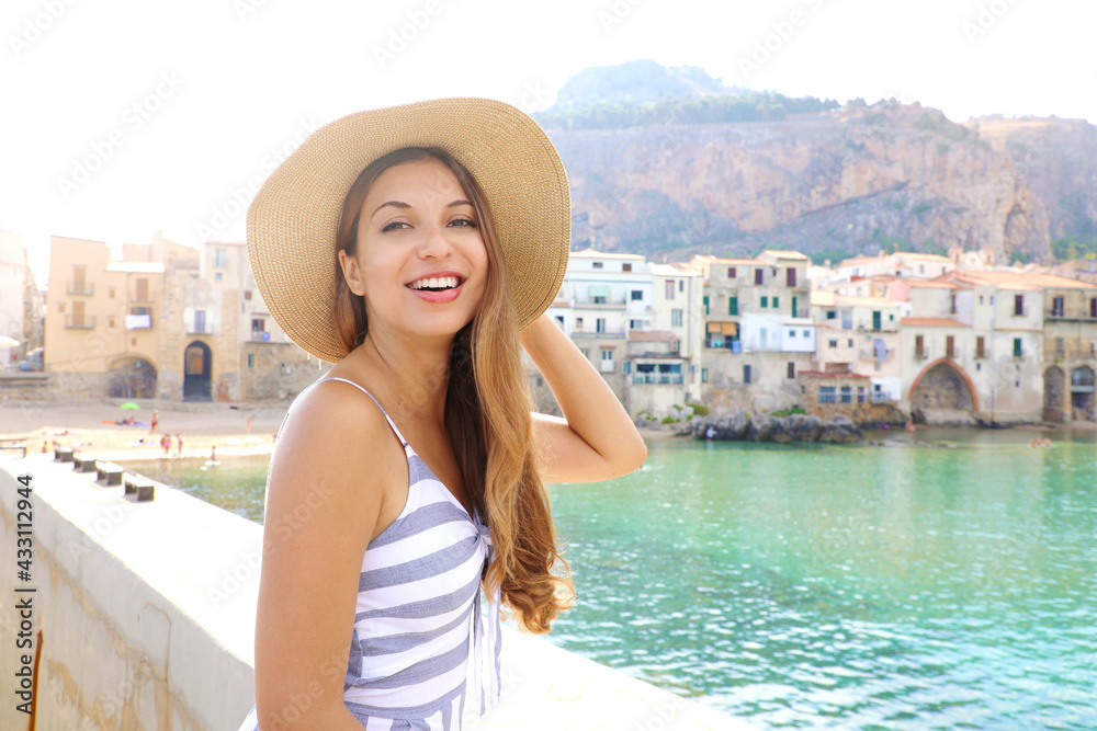 Pretty smiling woman with hat and summer dress looking at camera with Cefalu village on the background, Sicily, Italy