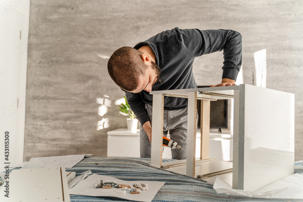 Obraz premium One Man with beard alone Putting Together Self Assembly Furniture at Home holding electric screwdriver screwing and putting together pieces - half length front view DIY concept real people copy space