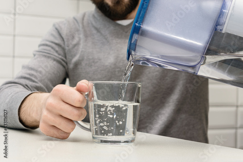 Man pours water from a filter jug into a glass in the kitchen, cropped image
