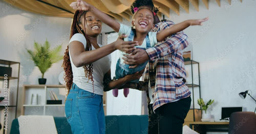 Cheerful african man and woman waving on hands their cute little daughter while staying at home. Young family in casual clothes spending free time actively.