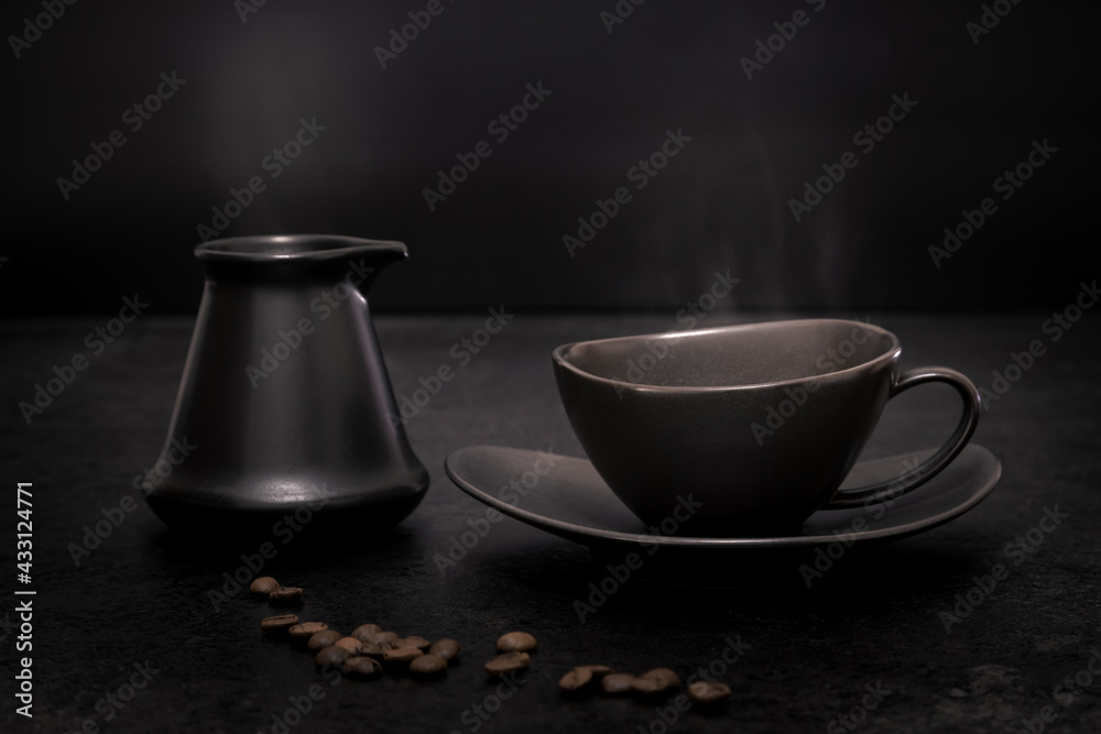 Low key still-life of a black coffee cup on a saucer and a black Turkish coffee pot with traces of steam above against a black background
