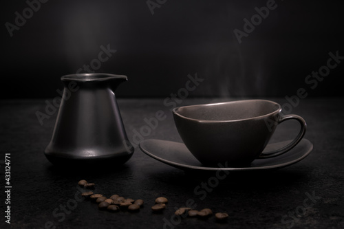 Low key still-life of a black coffee cup on a saucer and a black Turkish coffee pot with traces of steam above against a black background