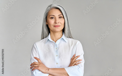 Stylish confident adult 50 years old Asian female psychologist standing arms crossed looking at camera at gray background. Portrait of sophisticated grey hair woman advertising products and services.