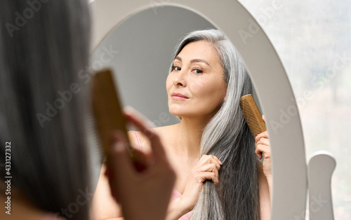 Obraz na plátně Senior attractive middle 50 years aged asian woman with gray hair looking at mirror reflection combing tangled gray hair