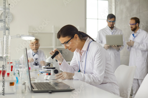 Virologist doing research studying in medical laboratory. Concentrated female virologist in goggles and latex gloves sits at table in laboratory and examines cells under microscope.