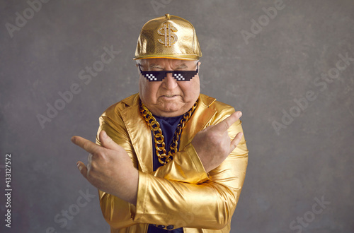 Retired older adult man in thug life glasses, gold chain and disco outfit isolated on gray background. Studio portrait senior pensioner in funny sunglasses looking at camera with angry face expression photo