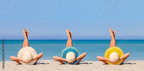 Fototapeta Naklejka Na Ścianę i Meble -  Three young girls on the beach wearing straw hats in the colors of the flag of Canary Islands. The concept of the perfect holiday in a Canary Islands resort. Focus on hats.