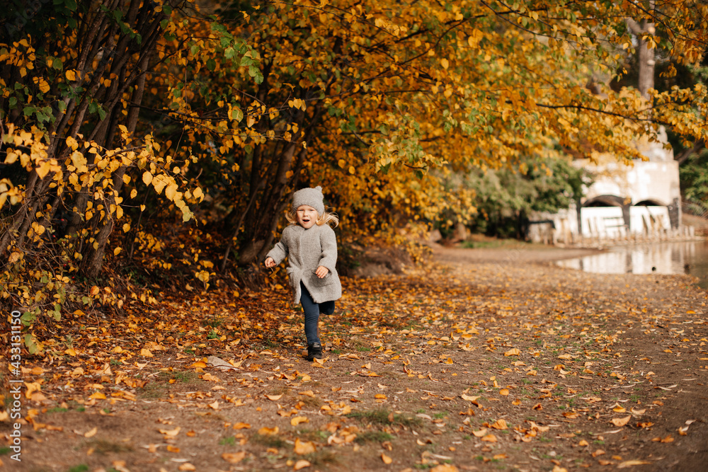 Fototapeta premium Goldener Herbst, ein 4-jähriges Mädchen in einem grauen Mantel läuft in der Nähe des Sees im Herbstpark