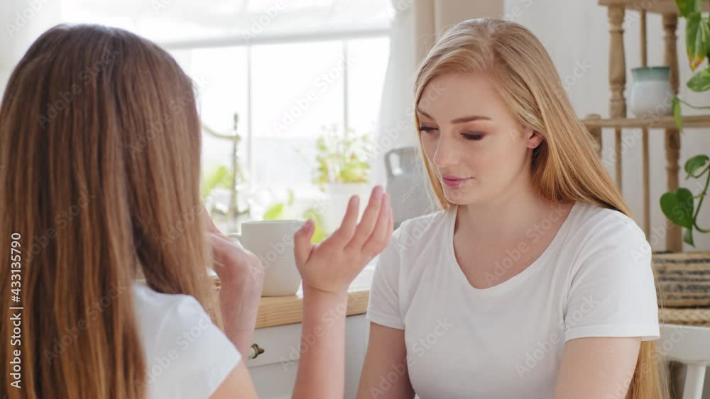 Indoors portrait adult beautiful blonde woman caring mother listens to unrecognizable teenage daughter child girl gesturing showing in air heart circle shape with hands sharing news sitting at home Indoors portrait adult beautiful blonde woman caring mother listens to unrecognizable teenage daughter child girl gesturing showing in air heart circle shape with hands sharing news sitting at home