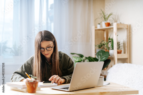 Thinking Freelance woman in eye glasses with pencil writing in note book, typing at laptop and working from home office. Happy girl on workplace at the desk. Distance learning online education.