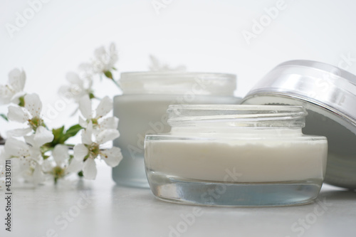 Beauty cream in glass jars on a light gray background. Decorated with white spring flowers. Unbranded skincare product. Cosmetic cream. Close up, selective focus, side view.