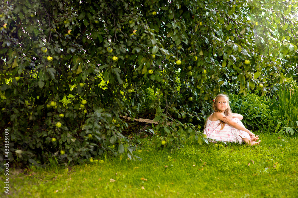 Blonde girl and toddler in light clothes sitting under an apple tree with green apples outdoors, healthy eating, healthy childhood, selective focus Blonde girl and toddler in light clothes sitting under an apple tree with green apples outdoors, healthy eating, healthy childhood, selective focus