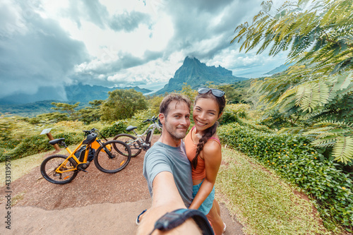 E-bike biking couple taking seflie on bikepacking travel vacation. Interracial tourists Asian woman, Caucasian man happy having fun on Moorea island ride, Tahiti, French Polynesia holiday adventure. photo