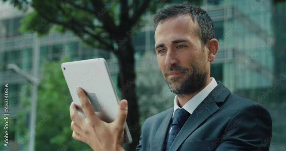 Cinematic shot of smiling businessman using tablet for sending emails and make calls for work in city park during break. Concept of technology, network, connection, corporate, finance, success. Cinematic shot of smiling businessman using tablet for sending emails and make calls for work in city park during break. Concept of technology, network, connection, corporate, finance, success.