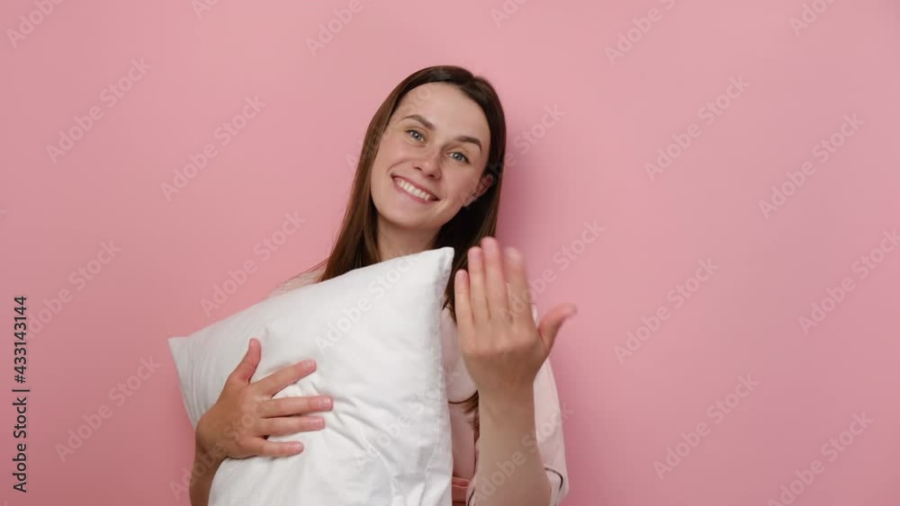 Attractive cute young European woman 20s old years in pajamas presenting and inviting to come with hand, hold white pillow, posing isolated on pink studio background. People emotion lifestyle concept Attractive cute young European woman 20s old years in pajamas presenting and inviting to come with hand, hold white pillow, posing isolated on pink studio background. People emotion lifestyle concept