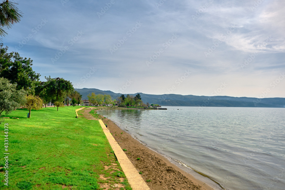 A shore and water's edge of  Nicaea (iznik) lake during sunny day and with beach covered by green trees and grass with small pathway near it and small hill background and blue sky.