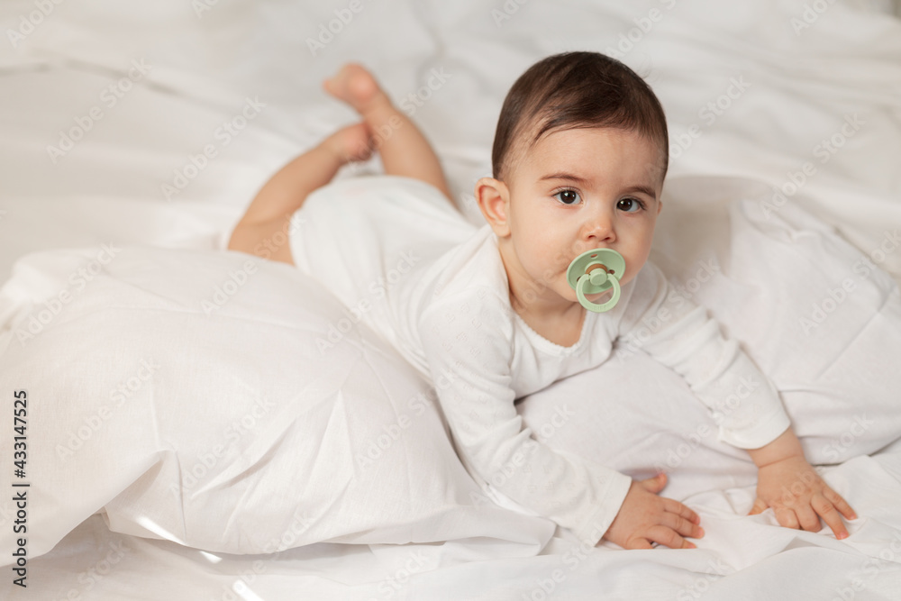 cute little mestizo girl lying in bed with a pacifier in her mouth in a white bodysuit with cotton bedding. High quality photo cute little mestizo girl lying in bed with a pacifier in her mouth in a white bodysuit with cotton bedding. High quality photo