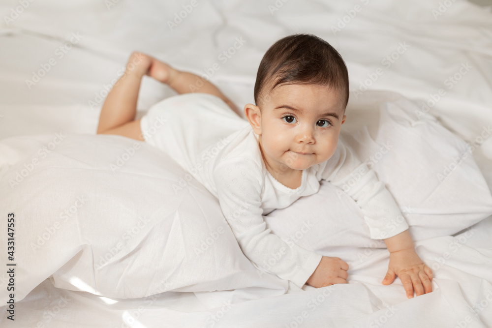 Cute little girl in a white bodysuit lies in bed with cotton linen. High quality photo. Cute little girl in a white bodysuit lies in bed with cotton linen. High quality photo.