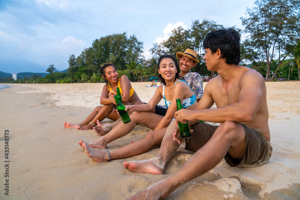 Fototapeta premium Group of Happy Asian man and woman friends sitting on the beach enjoy drinking beer with talking together at summer sunset. Male and female friendship relax and having fun outdoor lifestyle activity.