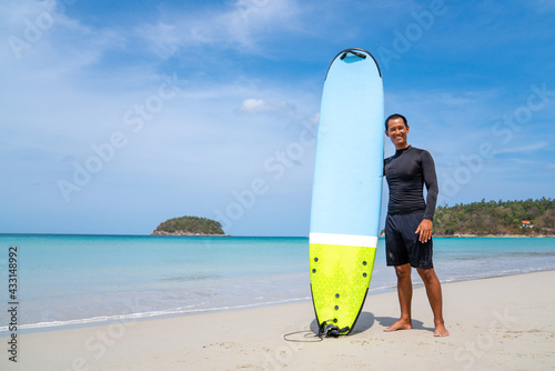 Canvas Print Attractive Asian man in wetsuit holding surfboard standing on tropical beach at summer sunny day