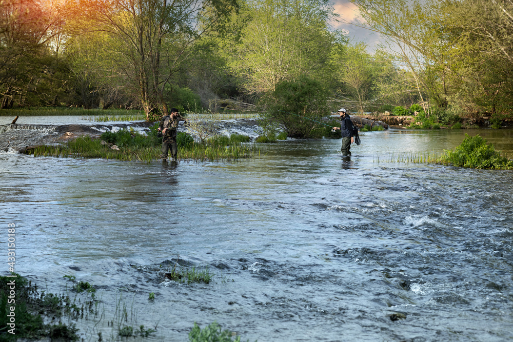 Two young teenagers in tall rubber fishing boots standing in the middle of the river while casting a fishing rod in the middle of a sunset space of copy Two young teenagers in tall rubber fishing boots standing in the middle of the river while casting a fishing rod in the middle of a sunset space of copy