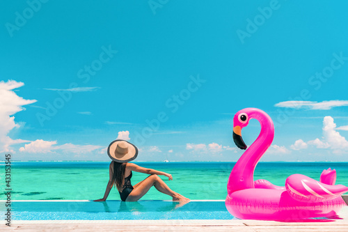 Vacation summer fun woman sunbathing with inflatable pink flamingo pool float by infinity swimming pool. Luxury travel holiday at overwater villa resort.