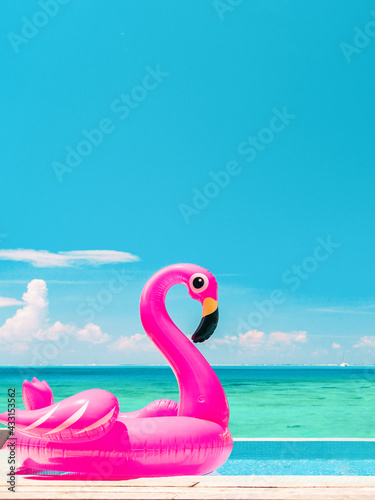 Summer vacation fun vertical crop of pink flamingo swimming pool toy float floating on infinity luxury resort pool with blue ocean background. © Maridav