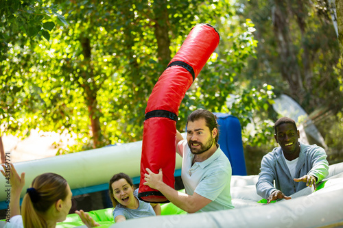 Adults having fun on inflatable amusement playground. Expressive bearded guy fighting off his friends with inflatable log while they trying to filch toy chickens © JackF