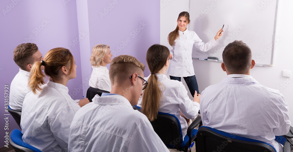 Obraz premium Girl medical student lecturing near whiteboard in front of teacher and group of students in auditorium.