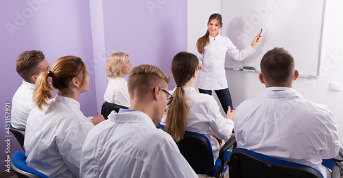 Girl medical student lecturing near whiteboard in front of teacher and group of students in auditorium.