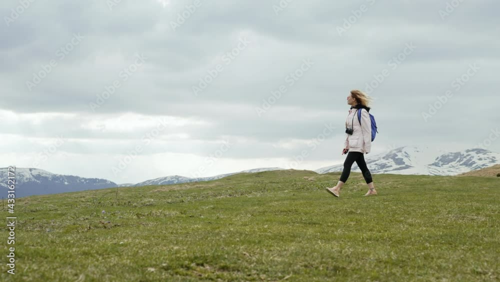Young traveller woman walking alone on valley of high mountain. Lady with backpack and camera. Snow-capped peaks of neighboring rocks. Slow motion. Young traveller woman walking alone on valley of high mountain. Lady with backpack and camera. Snow-capped peaks of neighboring rocks. Slow motion.