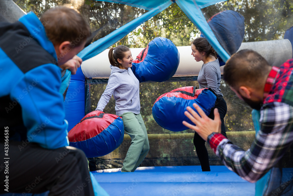 Obraz premium Friends having fun in outdoor amusement park, two men watching women in big boxing gloves boxing on inflatable ring