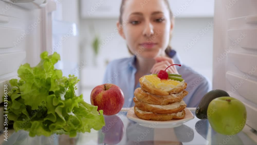 Beautiful young woman choosing between an apple and sweet cake. Healthy lifestyle. View from inside the working fridge. Beautiful young woman choosing between an apple and sweet cake. Healthy lifestyle. View from inside the working fridge.