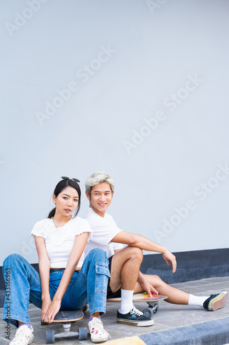 Asian handsome man and cute woman sitting on skateboards  looking straight  act smart  and smile in front of cool wall with copy space on wall in daylight time  summer holiday