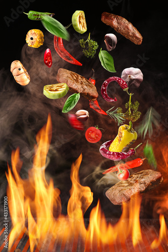 Flying beef stakes and vegetables above barbecue grill with fire on dark background