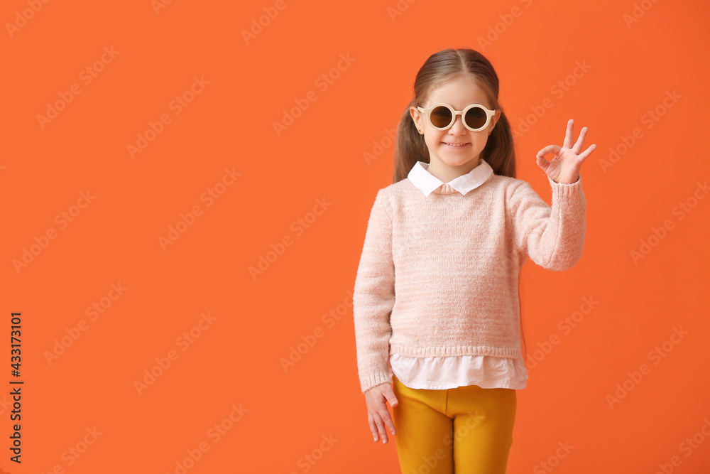Cute little girl wearing stylish sunglasses and showing OK on color background