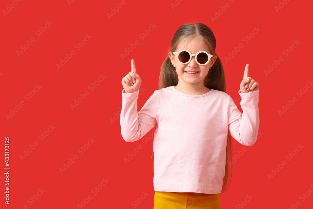 Cute little girl wearing stylish sunglasses and pointing at something on color background