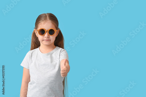 Cute little girl wearing stylish sunglasses and showing thumb-up on color background