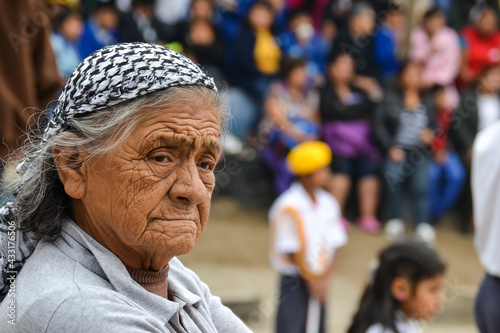 Beautiful grandmother with gray hair and a checkered scarf on her head, she has very marked wrinkles on her face and a sad look. blurred background of people who are not recognized.