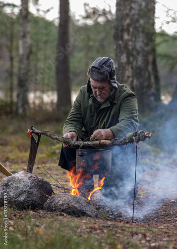 man in the forest trying to light a campfire, a tree branch with a pot over the campfire, blurred forest background, bonfire and smoke, autumn time