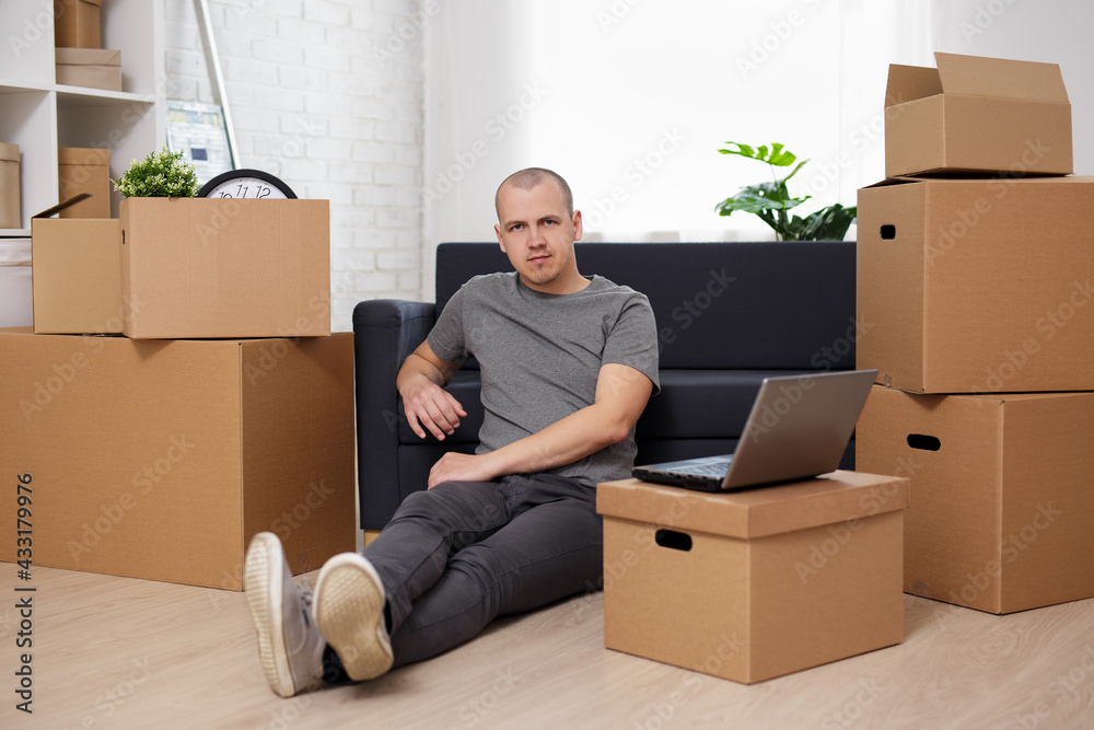 moving day concept - man sitting on floor with laptop and cardboard boxes in room after moving day moving day concept - man sitting on floor with laptop and cardboard boxes in room after moving day