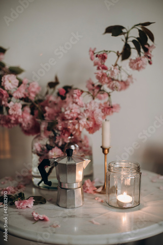 steel Italian Moka Pot on marble coffee table with pink flowers. burning candle in a transparent jar on a marble table with a vase of sakura branches.