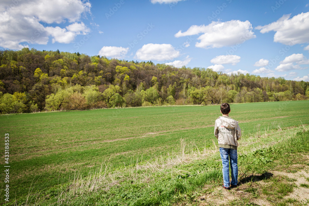 The boy stands in front of the field and the forest. Sunny summer day.