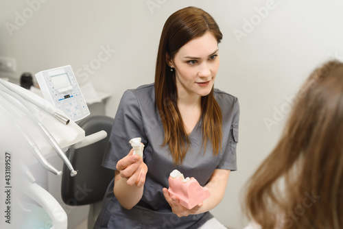 Female doctor talking to a young woman patient. Discussion of the treatment plan. The dentist shows a model of a dental implant. Modern dental clinic